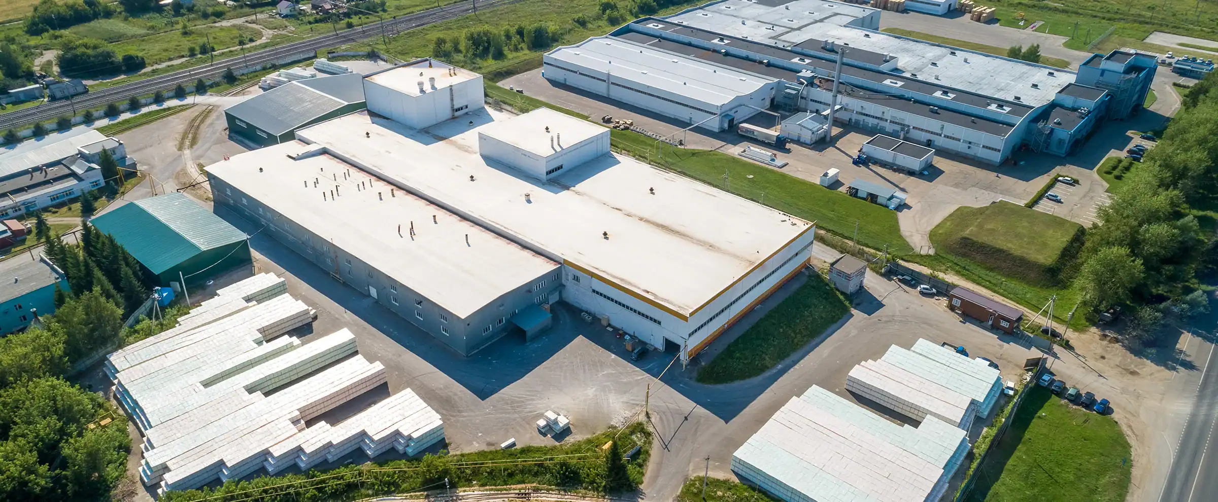 Flygbild över ett industriområde med stora fabriksbyggnader, lagerytor med staplat material och järnvägsspår i bakgrunden, omgivet av grönska.
