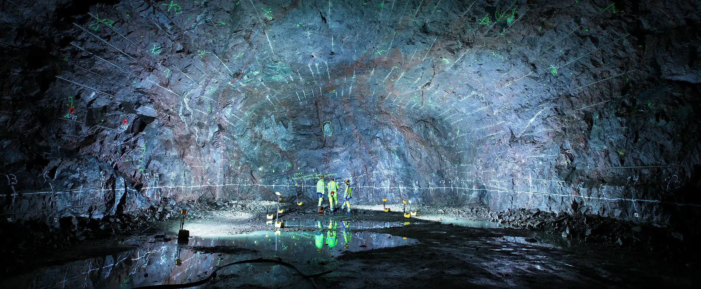Bergkartering i SKB:s Äspölaboratorium – tre personer i varselkläder undersöker en bergvägg markerad med mätlinjer och bergbultar i ett utsprängt bergrum.